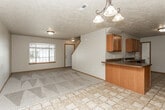 This two-story home is thoughtfully laid out across two levels, offering a functional and comfortable living environment. The main level presents an open-concept design where the kitchen flows seamlessly into the living and dining areas. The kitchen is well-equipped with warm wood cabinetry, dark countertops, tile flooring, and black appliances including a range, refrigerator, and built-in microwave. A peninsula with a sink serves as both a workspace and a visual divider between the kitchen and the adjacent carpeted living area. The living space is bright, benefiting from a large front-facing window that draws in natural light, and features a textured ceiling and a decorative pendant chandelier. A staircase leads to the upper level, where carpeted bedrooms offer a quiet retreat. The primary bedroom includes vaulted ceiling details, a pendant light fixture, and direct access to a private bathroom. Additional bedrooms are generously sized with natural light from windows. Bathrooms throughout the home are finished with tile flooring, wood vanities, and marble-style countertops, with the upper bath including a tub-shower combination.