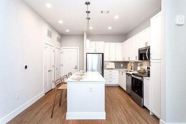 Cocina moderna con gabinetes blancos, iluminación bajo los gabinetes, protector contra salpicaduras de azulejos grises y encimeras de cuarzo blanco. - Avalon Carmel Commons