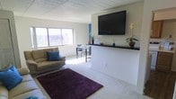 This single-floor residence offers a thoughtfully designed open-plan layout that seamlessly blends the living and dining areas into one cohesive space. The living area is anchored by a comfortable sofa accented with decorative pillows and a rich, deep-toned area rug that adds warmth and contrast to the light-colored carpeted flooring. A large flat-screen TV is mounted prominently on the wall above a sleek, dark ledge — a stylish divider that also serves as a display surface for tasteful greenery and décor accents. The dining area is positioned adjacent to the living space, bathed in natural light pouring through large windows that frame elevated views of the surrounding urban landscape. The neutral, light-toned walls and ceiling panels maintain a clean and airy atmosphere throughout. A pop of color from a blue accent wall adds a subtle visual focal point, enhancing the overall modern aesthetic. The space is compact yet efficiently arranged, making excellent use of its open floor plan to create a welcoming and functional environment suitable for both relaxation and entertaining.