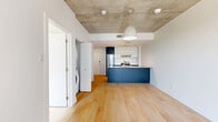 Located at 50 Place De L'Éclipse in Brossard, Quebec, this modern one-bedroom, one-bathroom unit offers a thoughtfully designed layout with a clean, contemporary aesthetic throughout. Light-toned hardwood flooring runs seamlessly across the living areas and bedroom, creating a warm and cohesive feel.

The open-concept kitchen is a standout feature, anchored by a bold navy-blue island with a light quartz countertop and an undermount sink. Stainless steel appliances, white upper cabinetry with dark accent uppers, and integrated pendant and track lighting give the kitchen a polished, functional character. The kitchen flows naturally into the main living area, making the space feel open and connected.

The bedroom is bright and compact, featuring a sliding double closet door for efficient storage and a window that frames elevated views of the surrounding urban landscape, including neighboring glass towers and green spaces below — a hallmark of a high-rise setting.

The bathroom is finished in a clean, all-white palette with large-format wall tiles, a soaker tub with a chrome shower fixture, wood-look floor tiles, and minimalist fixtures throughout.

Exposed concrete ceiling details in select areas add an industrial touch that complements the otherwise crisp, modern finishes. This unit is ideal for those seeking a well-appointed, efficient living space in a contemporary urban environment.