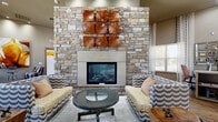 Located at 9701 Pearl St in Thornton, Colorado, this thoughtfully designed single-floor living space offers a welcoming and upscale communal environment. The room immediately impresses with its soaring ceilings, which lend an expansive, airy feel throughout. Rich, wide-plank hardwood flooring runs the length of the space, grounding the interior with warmth and texture.

A dramatic floor-to-ceiling natural stone fireplace serves as the room's central focal point, accented by bold metallic wall art in warm copper tones. Multiple seating arrangements are thoughtfully positioned around the space, including patterned lounge chairs and sofas, inviting both conversation and relaxation. A large communal dining table with colorful upholstered ottomans provides a functional gathering area bathed in natural light from oversized windows dressed in layered, patterned drapery.

A built-in bookshelf wall adds an intellectual and cozy library atmosphere to one side of the room, while a dedicated workstation with open shelving and task seating offers a productive nook. Recessed lighting throughout ensures the space is well-lit at all hours. The overall design blends contemporary and transitional styles, creating a multi-functional, vibrant, and inviting communal living room ideal for a variety of activities and gatherings.