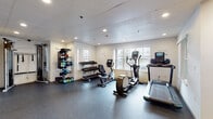 This fitness center is thoughtfully designed to accommodate a wide range of workout routines within a single-floor layout. The space is divided into distinct functional zones, each outfitted with purpose-specific equipment to support both cardio and strength training goals.

The cardio area features a treadmill, elliptical trainer, and a stationary recumbent bike, each equipped with integrated digital displays. Natural light filters in through multiple windows along the walls, creating a bright and energizing atmosphere enhanced by recessed ceiling lighting throughout.

The strength training section is anchored by a full power rack with barbell and weight plates, accompanied by a flat bench for pressing exercises. A row of spin bikes occupies an adjacent area, reflected in large wall-mounted mirrors that also help visually expand the space. A cable machine is available for functional and resistance-based training.

Free weights are neatly organized on a multi-tier dumbbell rack, while a separate storage unit holds kettlebells, resistance bands, foam rollers, and other accessories, keeping the floor space clear and organized.

A transitional entryway with light wood flooring connects to what appears to be an adjacent pool area through glass doors, with a water bottle filling station conveniently located nearby. The durable rubber flooring throughout the main workout zones provides both safety and comfort during exercise.