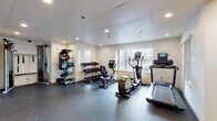 This fitness center is thoughtfully designed to accommodate a wide range of workout routines within a single-floor layout. The space is divided into distinct functional zones, each outfitted with purpose-specific equipment to support both cardio and strength training goals.

The cardio area features a treadmill, elliptical trainer, and a stationary recumbent bike, each equipped with integrated digital displays. Natural light filters in through multiple windows along the walls, creating a bright and energizing atmosphere enhanced by recessed ceiling lighting throughout.

The strength training section is anchored by a full power rack with barbell and weight plates, accompanied by a flat bench for pressing exercises. A row of spin bikes occupies an adjacent area, reflected in large wall-mounted mirrors that also help visually expand the space. A cable machine is available for functional and resistance-based training.

Free weights are neatly organized on a multi-tier dumbbell rack, while a separate storage unit holds kettlebells, resistance bands, foam rollers, and other accessories, keeping the floor space clear and organized.

A transitional entryway with light wood flooring connects to what appears to be an adjacent pool area through glass doors, with a water bottle filling station conveniently located nearby. The durable rubber flooring throughout the main workout zones provides both safety and comfort during exercise.