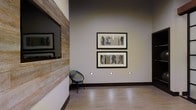 This single-story space showcases a thoughtfully designed interior that balances rustic textures with modern sensibilities. A striking reclaimed wood accent wall serves as a focal point, its weathered, natural grain providing a warm contrast against the smooth, neutral-toned walls. Embedded within this wooden feature is a large flat-screen display, seamlessly integrating technology into the organic material.

The open layout is anchored by light-toned flooring that reflects the ambient lighting, lending an airy and polished feel to the interior. Low-profile recessed step lights are installed along the base of the walls, adding a subtle, sophisticated glow that enhances the architectural lines of the space.

A curated gallery wall displays two framed artworks featuring figurative sketches, contributing a cultured and artistic atmosphere. A minimalist accent chair in muted teal is positioned nearby, offering a functional yet stylish seating option. Dark baseboards provide clean definition between the walls and floors, reinforcing the space's contemporary character. Overall, the space presents a refined, gallery-like ambiance.