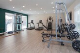 This fitness center is designed to accommodate a full range of workout routines within a clean, modern environment. The space features light hardwood-style flooring throughout, white walls accented by a bold teal feature wall, and recessed ceiling lighting that ensures the entire area is well-lit and welcoming.

The cardio zone is equipped with treadmills, stair climbers, and elliptical machines, all positioned near windows fitted with horizontal blinds that allow natural light to filter in. A wall-mounted flat-screen television provides entertainment during cardio sessions. Adjacent to this area, a dedicated spin zone houses multiple stationary bikes with integrated digital displays.

The strength training area is equally comprehensive, featuring a cable and pulley machine, adjustable weight benches, a full rack of dumbbells in varying weights, a medicine ball rack stocked with an assortment of weighted balls, and a barbell plate storage rack. A large mirrored wall in one section of the space enhances the sense of openness and allows for form checks during exercise.

Large windows along multiple walls bring in ample natural light and offer views of the surrounding outdoor area, contributing to an energizing atmosphere. The layout is thoughtfully organized, providing clear zones for cardio, strength, and functional training while maintaining generous open floor space for movement and stretching.