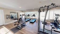 Located at 1167 Janaf Pl in Norfolk, Virginia, this fitness center offers a thoughtfully designed workout facility on a single floor. The space is finished with durable wood-style flooring, crown molding, and recessed lighting, creating a clean and inviting atmosphere conducive to exercise.

Large windows line multiple walls of the facility, flooding the interior with abundant natural light and providing open views of the surrounding greenery and outdoor areas. A glass door provides direct access to an outdoor patio, further enhancing the connection between the interior and exterior environment.

The fitness center is well-equipped with a comprehensive selection of cardio and strength training equipment, including multiple treadmills, elliptical machines, a rowing machine, and various cable and resistance strength machines with adjustable benches. Full-length mirrors are mounted along one wall to assist users with form and technique while also visually expanding the space.

A compact kitchenette is integrated into one corner of the facility, complete with dark cabinetry, a stainless steel refrigerator, and a microwave, offering added convenience for users. The neutral-toned walls and open layout contribute to a bright, airy ambiance that makes the space feel welcoming and functional for a variety of fitness routines.