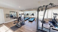 Located at 1167 Janaf Pl in Norfolk, Virginia, this fitness center offers a thoughtfully designed workout facility on a single floor. The space is finished with durable wood-style flooring, crown molding, and recessed lighting, creating a clean and inviting atmosphere conducive to exercise.

Large windows line multiple walls of the facility, flooding the interior with abundant natural light and providing open views of the surrounding greenery and outdoor areas. A glass door provides direct access to an outdoor patio, further enhancing the connection between the interior and exterior environment.

The fitness center is well-equipped with a comprehensive selection of cardio and strength training equipment, including multiple treadmills, elliptical machines, a rowing machine, and various cable and resistance strength machines with adjustable benches. Full-length mirrors are mounted along one wall to assist users with form and technique while also visually expanding the space.

A compact kitchenette is integrated into one corner of the facility, complete with dark cabinetry, a stainless steel refrigerator, and a microwave, offering added convenience for users. The neutral-toned walls and open layout contribute to a bright, airy ambiance that makes the space feel welcoming and functional for a variety of fitness routines.