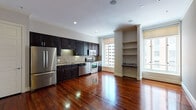 This single-floor residence offers a thoughtfully designed layout that balances modern finishes with classic architectural details. The open-concept kitchen and living area serves as the heart of the space, showcasing rich, deeply stained hardwood floors that gleam with a polished finish. The kitchen is fully equipped with a complete suite of stainless steel appliances, including a French door refrigerator, electric range with oven, built-in microwave, and dishwasher. Dark espresso cabinetry, some featuring glass-front doors, lines the walls and is complemented by granite countertops and a distinctive striped tile backsplash. Large windows flood the area with natural light while offering urban cityscape views, and a built-in shelving unit with a granite ledge adds both character and functionality to the living zone. Crown molding accents the ceilings throughout, lending an air of refinement. The bathroom is finished with warm travertine-style tile flooring and wall surround, a walk-in glass-enclosed shower, a dark vanity with granite countertop, and a lighted mirror. The bedroom features plush neutral carpeting and a clean, understated palette, providing a calm and restful retreat. The overall ambiance of the space is urban and polished, blending contemporary conveniences with timeless design elements.