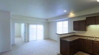This single-floor residential space offers a clean, functional layout designed for comfortable everyday living. The open living area is bathed in natural light, courtesy of large sliding glass doors fitted with vertical blinds that lead to an outdoor area, as well as a classic grid-pane window that further brightens the interior. The space features plush wall-to-wall carpeting that adds warmth and comfort underfoot, while the textured ceiling with recessed lighting provides a practical yet polished overhead finish. A kitchen pass-through bar with rich, dark wood cabinetry serves as a natural divider between the kitchen and living areas, offering both a visual anchor and added counter space. The neutral color palette of soft whites and warm beiges throughout creates an airy, inviting atmosphere that is easy to personalize. The overall ambiance is light, open, and welcoming, with a straightforward design that maximizes the sense of space. The combination of ample natural light, functional cabinetry, and open floor planning makes this a versatile and practical living environment suitable for a wide range of occupants.