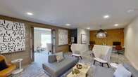 This single-floor workspace offers a sophisticated blend of modern design and warm, natural materials across several well-defined functional zones. The main lounge area serves as the heart of the space, featuring a contemporary gray sectional sofa, a low-profile coffee table with decorative greenery, and a patterned area rug. Sleek white accent chairs provide additional seating, while bold typographic wall art and a textured wood accent wall add visual character. Recessed lighting throughout ensures a clean, well-lit environment.

A dedicated co-working area is anchored by a statement woven pendant light and features a workstation desk, monitor, and ergonomic seating against a rich, textured amber wall panel. A compact round dining or breakout table nearby, also topped by a striking woven pendant, offers an intimate setting for small group discussions.

The conference room is a standout feature, centered around a stunning live-edge wood slab table paired with leather chairs, a wall-mounted display, a multi-globe chandelier, and a vibrant mosaic artwork. Natural light flows in through a large window with outdoor views of landscaped grounds.

A unique relaxation nook showcases a built-in herringbone wood daybed alcove flanked by wall sconces and white lounge chairs. Private rooms accessible via frosted glass-panel doors offer additional enclosed meeting or focus spaces, completing this versatile and design-forward environment.