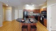 This single-floor residence offers a thoughtfully designed open-concept layout that seamlessly connects the kitchen, dining, and living areas. The kitchen is a functional centerpiece, featuring rich wood cabinetry, dark granite countertops, black appliances including a refrigerator, stove, and built-in microwave, and a curved island with bar seating illuminated by track lighting above. Hardwood-style flooring extends throughout the main living area, which is bright and welcoming thanks to large windows and a glass door that fills the space with natural light.

The living room provides a comfortable, open setting with room for multiple seating arrangements. The bedrooms are carpeted for warmth and comfort, each featuring windows that bring in natural light and closet storage. One bedroom is well-suited as a home office or flex space. The primary bedroom includes access to a walk-in closet area.

Two bathrooms serve the residence — one featuring a glass-enclosed shower and the other a full soaking tub with shower combination and a white vanity with ample storage. Neutral warm tones throughout create a cohesive and inviting ambiance across all rooms.