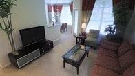 This single-floor residence offers a well-designed open-concept layout that seamlessly connects the living and dining areas. The living room is appointed with plush carpeting, a sleek black media console supporting a flat-screen television, and a glass-top coffee table that adds a contemporary touch. A cushioned accent chair and a sofa provide comfortable seating, while a side table with a lamp offers warm ambient lighting. Bold red and sage green two-tone curtains frame the windows, which allow generous natural light to fill the space. A pass-through opening connects the living area to the dining space, which features a chandelier overhead and is furnished with a dining table and upholstered chairs in a rich red tone. Additional windows fitted with horizontal blinds further brighten the interiors. The neutral, light-toned walls and carpeted floors create a cohesive, understated backdrop that allows the furnishings and drapery to stand out. Decorative accents, including framed photos on the side table, contribute to the warm and inviting atmosphere throughout this thoughtfully arranged single-level residence.