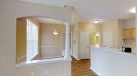 This single-floor home offers a thoughtfully designed open-concept layout that seamlessly connects the primary living areas. The space is characterized by warm beige and neutral wall tones that create a welcoming and bright ambiance throughout.

A standout architectural feature is the decorative white column framing the arched entryway that divides the dining and living areas, adding a classic, elegant touch to the interior. The hardwood flooring flows consistently through the main areas, lending warmth and continuity to the space.

The kitchen features a raised breakfast bar countertop, providing both a functional workspace and a casual dining option. Natural light enters through white-shuttered windows, brightening the living and dining areas. A pendant light fixture hangs in the dining zone, complementing the flush-mount ceiling lighting used throughout.

Interior doors with raised paneling lead to additional rooms, including what appears to be a bathroom with visible vanity lighting. The layout provides a practical flow between spaces, making everyday movement through the home comfortable and intuitive.

Overall, this home combines classic design elements — arched doorways, decorative columns, and clean white trim — with functional, open living spaces to create a warm and inviting environment.