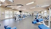 This spacious, single-floor fitness center is thoughtfully designed to support a full range of workout routines. The open layout accommodates a generous collection of white-framed strength training machines with blue upholstery, including cable pulley systems, leg press and extension machines, chest and shoulder press stations, and adjustable benches. The cardio section features multiple treadmills positioned along a windowed wall, allowing exercisers to enjoy natural light and views of the surrounding outdoor area. Full-length mirrors line a portion of the walls, visually expanding the space and providing form feedback during workouts. The flooring throughout is a durable gray exercise surface suited for high-traffic use. Overhead, flush-mounted fluorescent light panels provide consistent, even illumination, while multiple ceiling fans contribute to a comfortable training environment. French doors along one wall open directly to an outdoor pool area, enhancing ventilation and providing a seamless indoor-outdoor connection. A first aid station is mounted near the exit for safety. The overall aesthetic is clean and functional, with a neutral warm-beige color palette that creates a bright and welcoming atmosphere for fitness activities.