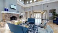 This single-floor common area presents an impressive and well-appointed lounge space defined by its dramatic double-height ceilings and an abundance of natural light. Clerestory windows positioned high along the upper walls flood the interior with daylight, enhancing the open, airy atmosphere throughout. The focal point of the room is a stately stone fireplace with a classic white mantle, flanked by a decorative console and greenery, adding warmth and character to the space. A wall-mounted television above the fireplace adds a functional element to the elegant setting.

The seating area is thoughtfully arranged around a glass-and-gold coffee table, featuring a navy blue sofa, coordinating patterned armchairs, and chartreuse accent ottomans, all anchored by a blue geometric area rug. Wall sconces with ornate detailing provide ambient lighting along the neutral gray walls. French doors at the far end of the room invite natural light from outside and offer views of an exterior space. The light tile flooring throughout ties the design together, creating a cohesive, sophisticated, and inviting environment.