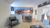 This single-floor residence showcases a thoughtfully designed open-concept layout that seamlessly connects the kitchen, dining, and living areas into one cohesive and functional space. The kitchen is equipped with rich, warm-toned cabinetry that extends to the upper walls, providing generous storage, and is complemented by modern built-in appliances, including a microwave and refrigerator. A breakfast bar peninsula serves as a natural divider between the kitchen and the rest of the living space, adding both utility and visual structure to the layout. An accent wall in a deep steel blue anchors the kitchen and dining area, adding a bold yet sophisticated pop of color that contrasts elegantly with the warm wood tones. The dining area features a classic dark-framed table with coordinating chairs, illuminated by a multi-light chandelier that provides warm, focused lighting. Hardwood flooring extends through the kitchen and dining areas, transitioning into plush carpet in the living room, which enhances comfort underfoot. The living area is accented with stylish furnishings, including a patterned settee, contributing to an inviting and curated ambiance. The space benefits from a clean, bright atmosphere thanks to white ceilings and neutral walls throughout.