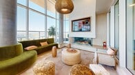 Located at 110 Beech Street in San Diego's vibrant Little Italy neighborhood, this single-floor living space offers a refined and visually striking interior defined by thoughtful architectural design and premium finishes throughout. Soaring double-height ceilings amplify the sense of openness, while an expansive wraparound curtain wall of floor-to-ceiling glass floods the space with abundant natural light and frames panoramic views of the San Diego skyline, waterfront, and outdoor terrace with a pool beyond.

The living area is anchored by a sleek, modern fireplace integrated into a fluted wall panel beneath a wall-mounted screen, complemented by plush velvet seating in earthy olive tones and woven accent ottomans. Adjacent lounge nooks feature deep sectional sofas set against sheer linen curtains, with the cityscape as a backdrop. Structural concrete columns add an industrial counterpoint to the warm wood and marble accents found throughout.

A curved central bar serves as a focal point, clad in geometric patterned tile and topped with a white stone surface, surrounded by wooden barstools with teal upholstery. Orb pendant lighting cascades from a sculpted curved ceiling detail overhead, lending a dramatic, curated ambiance. Lush tropical greenery and sculptural décor pieces complete the sophisticated, resort-inspired aesthetic of this exceptional San Diego space.