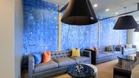 This single-floor space makes a strong visual impression through its bold, curated design choices. The most commanding feature is the floor-to-ceiling abstract mural spanning the walls, rendered in rich shades of cobalt and electric blue with textured, gestural brushwork evoking a forest or natural landscape. The mural creates an immersive, art-forward atmosphere that anchors the entire room.

The seating area is composed of contemporary tufted gray sofas and armchairs arranged around a sleek dark marble-topped cocktail table with a sculptural chrome accent piece at its center. Colorful throw pillows in coral, blue, and yellow add vibrant contrast against the neutral upholstery, contributing to a lively yet sophisticated palette.

Overhead, a dramatic oversized black cone pendant light commands attention, serving as a bold architectural statement that draws the eye upward. Recessed ceiling lighting supplements the pendant, ensuring the space is well-illuminated throughout. The overall ambiance is modern and design-driven, balancing artistic expression with comfortable, lounge-style functionality.