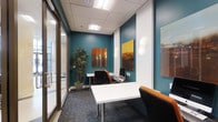 This single-floor office space combines functionality with contemporary design to create a polished and inspiring work environment. The interior is defined by striking teal accent walls that lend a bold, energetic character to the space, contrasted against clean white surfaces and a drop ceiling fitted with bright, recessed panel lighting that ensures consistent, even illumination throughout.

The workspace is thoughtfully furnished with sleek white desks and modern ergonomic chairs featuring distinctive orange upholstery, adding a vibrant pop of color that complements the overall design palette. Patterned carpet flooring in deep, muted tones grounds the space with a sense of sophistication while offering visual texture underfoot.

Curated wall art in warm amber and earthy tones adorns the teal walls, contributing to an engaging and visually dynamic atmosphere. Globe-shaped wall sconces provide additional ambient lighting and serve as tasteful decorative elements. A tall indoor plant adds a refreshing natural touch, softening the crisp modern aesthetic.

A glass partition wall along one side allows for an open, connected feel between areas while maintaining a sense of structure and privacy. Overall, the space strikes a refined balance between professional utility and contemporary design sensibility.