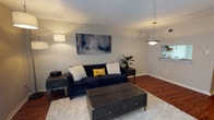 This two-story space combines modern design with a warm, inviting ambiance. The living area showcases rich hardwood flooring that anchors the room, complemented by a large, plush white area rug that adds texture and comfort underfoot. A deep navy blue sofa serves as the focal point, styled with contrasting white faux-fur and yellow accent pillows, creating a bold yet approachable aesthetic. A rustic dark wood coffee table with drawer storage sits at the center, balancing the softness of the rug with a grounded, substantial feel. Matching dark side tables flank the sofa, providing functional surface space. Wall art above the seating area features an abstract canvas in navy, grey, and blush tones, tying the color palette together cohesively. Layered lighting includes a sleek floor lamp and a geometric pendant light near the transitional space leading to an adjacent area, likely a kitchen or dining space, visible in the background. The neutral grey walls and white ceilings keep the space feeling open and bright, while the overall design reflects a curated, contemporary style carried throughout both floors.