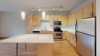 Located at 3900 Excelsior Blvd in St. Louis Park, Minnesota, this single-floor residence offers a thoughtfully designed layout that balances functionality with contemporary style. The kitchen is a standout feature, showcasing light maple cabinetry, white countertops, a classic subway tile backsplash, and a full suite of stainless steel appliances including a range, microwave, dishwasher, and refrigerator. A central island with a sleek white surface and pendant lighting above provides additional prep space and casual dining potential. Track lighting runs along the ceiling, ensuring the kitchen is well-lit throughout. Light hardwood flooring flows through the kitchen and living areas, creating a warm and cohesive feel. The open layout connects the kitchen seamlessly to the living space, which benefits from a sliding glass door leading to a private balcony with views of surrounding greenery. The bedroom is a comfortable retreat, featuring plush carpeting and a set of windows that invite natural light and outdoor views. The full bathroom is finished with neutral tile flooring, a soaking tub with shower, a light wood vanity, and clean white walls that contribute to a bright, airy atmosphere. The overall design is clean and modern, with warm wood tones throughout.