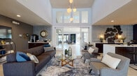 Located at Feather Sound Circle East in Clearwater, Florida, this single-story community clubhouse offers a thoughtfully designed gathering space with a contemporary aesthetic. The main lounge area immediately impresses with soaring double-height ceilings, clerestory windows that flood the space with natural light, and a striking pendant light fixture. Light-toned large-format tile flooring runs throughout, complementing the neutral and bold navy color palette.

The open-concept layout connects the lounge seating area — furnished with charcoal sofas, a glass-top coffee table, and layered area rugs — to a secondary lounge featuring a large wall-mounted television, decorative circular wall art, and a media console. A coffee bar and kitchenette area is outfitted with rich dark wood cabinetry, vibrant blue tile backsplash accents, and a curved white countertop island with bar seating, ideal for casual gatherings.

A sunlit dining nook, framed by a deep navy accent wall and a bank of large windows, offers direct views of the outdoor pool, palm trees, and waterway beyond — creating a bright, resort-like atmosphere. Frosted glass interior doors and structural columns add architectural interest throughout. The space is well-suited for resident events, social gatherings, or everyday use.