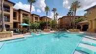 Pool 2 is a beautifully appointed outdoor aquatic retreat that embodies a resort-style atmosphere. The centerpiece is a generously sized swimming pool with crystal-clear turquoise water, designed to invite relaxation and leisure. The pool deck is finished in a light-toned stone surround, providing ample space for sunbathing and socializing.

Surrounding the pool, a curated selection of aqua-colored lounge chairs and chaise lounges offer comfortable spots for unwinding, complemented by shaded seating areas covered by teal market umbrellas for those seeking relief from the sun. A pergola structure anchors one end of the pool area, adding architectural interest and additional shaded gathering space.

Tall, mature palm trees are strategically placed throughout the area, lending a tropical ambiance and a sense of established greenery. The backdrop features warm, earth-toned Mediterranean-style buildings with balconies and arched architectural detailing, creating an enclosed and private feel. Open skies with drifting clouds above complete the inviting, vacation-inspired environment, making this pool area a standout outdoor amenity.