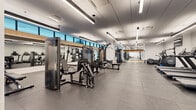 This contemporary fitness center is thoughtfully designed to accommodate a full spectrum of workout needs across a single, expansive floor. The space is organized into distinct functional zones, each outfitted with professional-grade equipment. The cardio section features multiple treadmills, elliptical machines, a stair climber, and a rowing machine, all positioned for ease of use and accompanied by a wall-mounted television for entertainment. The strength training area is comprehensively equipped with a variety of cable machines, multi-functional weight stations, a dedicated dumbbell rack, and free weight benches, catering to both beginners and advanced users. A separate open-floor studio zone, finished with light hardwood flooring, provides ample space for stretching, yoga, cycling, or group fitness activities, and includes accessories such as dumbbells and fitness mats. Throughout the facility, large floor-to-ceiling windows along one wall flood the space with natural light and offer views of the surrounding streetscape. The interior is finished with large-format gray tile flooring, crisp white walls, and a clean dropped ceiling fitted with linear LED lighting, creating a bright, modern, and motivating environment. The overall design balances functionality with a polished aesthetic suited to a premium fitness facility.