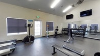 This single-floor fitness center offers a thoughtfully designed workout space suited for a variety of exercise routines. The room features vaulted ceilings that lend an open, airy feel, while large windows fitted with blackout roller shades allow for adjustable natural light throughout the day. Durable dark rubber flooring covers the workout area, providing both safety and functionality.

The space is outfitted with a range of equipment to support diverse training needs. A dumbbell rack lines one wall, stocked with multiple weight increments, while a weight bench and barbell setup offer options for strength training. A stationary upright bike provides cardio capability. A full-length wall mirror enhances the sense of space while allowing users to monitor their form during workouts. A wall-mounted flat-screen television adds an entertainment element to training sessions.

Bright fluorescent lighting ensures the room remains well-lit at all times, complementing the natural light from the windows. A dedicated exit door provides convenient access to and from the facility. Posted fitness center rules reflect an organized and maintained environment. Overall, this fitness center delivers a clean, functional, and comfortable setting for residents or members seeking a reliable on-site exercise facility.