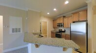 This single-floor residence offers a well-designed open-concept layout that seamlessly connects the kitchen and living areas. The kitchen is a standout feature, showcasing warm honey-toned cabinetry, granite countertops with a subtle speckled pattern, and a full suite of stainless steel appliances including a gas range, microwave, and refrigerator. A peninsula with an extended breakfast bar provides both additional prep space and casual seating, making it ideal for everyday dining and entertaining. The kitchen also includes an undermount sink and is finished with tile flooring that transitions smoothly into the adjacent spaces. Crown molding adds a refined architectural detail throughout, while recessed lighting illuminates the space with a clean, modern feel. The living area features plush carpet flooring, contributing to a comfortable and inviting atmosphere. Neutral wall tones throughout create a versatile backdrop suitable for a variety of interior styles. An interior door visible from the main living space leads to additional areas of the home, suggesting a functional and well-organized floor plan. Overall, the space presents a cohesive and move-in ready environment with quality finishes and thoughtful design details.