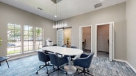 This single-floor professional workspace is thoughtfully designed to accommodate a variety of work styles and collaborative needs. The centerpiece of the space is a well-appointed conference room featuring soaring ceilings and a striking cluster of teardrop pendant lights suspended above a large oval white table surrounded by quilted navy blue rolling chairs. A wall of multi-pane windows floods the room with natural light and offers views of the surrounding greenery, while patterned blue-gray carpet adds a modern, textural element underfoot. A sleek barn-style sliding door with frosted glass panels leads to an adjacent area, maintaining an open yet defined flow throughout the space.

A private office nook, accessible through the barn door, provides a more intimate setting with a shiplap accent wall and a dedicated desk and chair, ideal for focused individual work. A secondary private room features a bold navy accent wall contrasted against whitewashed shiplap, complete with a built-in desk and ample power outlets. A cozy lounge corner with a round pedestal table, a sculptural wall clock mounted on shiplap, and plush seating offers a relaxed alternative for informal meetings or brief respites. Throughout, neutral gray walls, contemporary artwork, and curated greenery contribute to a polished, professional ambiance.