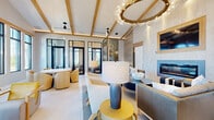 This elegantly designed single-floor space offers a generous layout encompassing a grand entrance, living room, dining area, and additional lounge space. The entrance makes an immediate impression with soaring ceilings, large-format hexagonal tile flooring, floor-to-ceiling black-framed windows, and a striking hand-painted ceiling mural. Bold large-scale artwork and gold-accented seating set a refined tone from the moment of arrival.

The living area continues the sophisticated aesthetic with exposed wood ceiling beams, a dramatic circular chandelier, a sleek linear fireplace set against a textured stone wall, and curated seating arrangements that invite conversation and relaxation. Black-framed glass partitions create visual separation while maintaining an open, airy feel throughout.

The dining space is anchored by a substantial live-edge communal table paired with woven bar-height chairs, illuminated by geometric black-iron pendant chandeliers. Open shelving, a beverage station, and outdoor-facing windows flood the area with natural light and views of surrounding greenery.

Throughout the space, a cohesive coastal-contemporary palette of warm whites, natural wood tones, and accent yellows creates a welcoming yet upscale ambiance. Multiple access points lead to outdoor areas, further enhancing the connection between interior comfort and the surrounding environment.