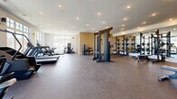 Located at 3271 Hilltown Dr in Kelowna, BC, this fitness facility offers a generously sized, single-floor exercise space designed to support a comprehensive workout experience. The main exercise room is bright and airy, benefiting from an abundance of natural light streaming through large, multi-pane windows that frame views of the surrounding Kelowna landscape, including mature trees and an outdoor balcony area accessible via full-height black-framed glass doors. The interior is finished with clean white ceilings fitted with recessed pot lighting, while the flooring transitions between light hardwood-style surfaces and durable rubber flooring in the workout zones. The facility is thoughtfully organized into distinct training areas. A dedicated free weights section features an extensive dumbbell rack, flat benches, and kettlebells, all set against a full-length mirror wall framed by warm wood slat paneling. The strength zone includes multi-functional cable machines and squat racks. The cardio area is lined with treadmills, elliptical trainers, and additional cardio equipment, all positioned to take advantage of the outdoor views. Two hallways connect and service the space. The overall ambiance is modern, clean, and professionally outfitted, making it a well-rounded fitness destination.