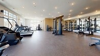 Located at 3271 Hilltown Dr in Kelowna, BC, this fitness facility offers a generously sized, single-floor exercise space designed to support a comprehensive workout experience. The main exercise room is bright and airy, benefiting from an abundance of natural light streaming through large, multi-pane windows that frame views of the surrounding Kelowna landscape, including mature trees and an outdoor balcony area accessible via full-height black-framed glass doors. The interior is finished with clean white ceilings fitted with recessed pot lighting, while the flooring transitions between light hardwood-style surfaces and durable rubber flooring in the workout zones. The facility is thoughtfully organized into distinct training areas. A dedicated free weights section features an extensive dumbbell rack, flat benches, and kettlebells, all set against a full-length mirror wall framed by warm wood slat paneling. The strength zone includes multi-functional cable machines and squat racks. The cardio area is lined with treadmills, elliptical trainers, and additional cardio equipment, all positioned to take advantage of the outdoor views. Two hallways connect and service the space. The overall ambiance is modern, clean, and professionally outfitted, making it a well-rounded fitness destination.