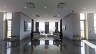 This sophisticated single-floor space is defined by its clean, contemporary design and refined aesthetic. The main living area immediately impresses with its expansive floor-to-ceiling windows that flood the interior with natural light while framing sweeping city skyline views. Dark, floor-length curtains flank the windows, adding a dramatic contrast to the otherwise light and neutral color palette. Underfoot, large-format polished tiles in a cool grey tone reflect light beautifully, enhancing the sense of spaciousness throughout. The living area is thoughtfully arranged with plush, modern seating — including cushioned armchairs and a central coffee table — creating an inviting yet elegant atmosphere. A statement chandelier hangs at the room's center, serving as a focal point that bridges classic glamour with modern minimalism. Recessed lighting throughout the ceiling provides balanced ambient illumination, complementing the natural light. A large flat-screen television is mounted flush against one wall, while curated artwork adds visual interest. The overall ambiance is one of understated luxury — a meticulously designed space that balances comfort, style, and a strong architectural presence.