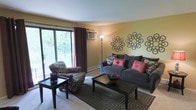 This single-floor space centers around a well-appointed living room that blends comfort and style. The room is bathed in natural light streaming through a large sliding glass door, which opens to a balcony overlooking a lush, tree-lined view — a standout feature that brings the outdoors in. Dark burgundy curtains frame the door, adding a rich contrast to the warm yellow-toned walls. A wall-mounted air conditioning unit ensures year-round comfort, while a baseboard heater runs along the exterior wall.

The living area is furnished with a plush blue-grey sofa accented with pink and patterned throw pillows, complemented by a coordinating patterned armchair. Two dark wood coffee and side tables ground the seating arrangement, while a geometric area rug adds texture and definition to the carpeted floor. Decorative circular metal wall art in a floral pattern serves as a focal point on the main wall, adding an artistic touch. A tall floor lamp provides warm ambient lighting, contributing to the overall inviting and lived-in atmosphere of the space.