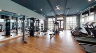 Located at 7111 Rock Fish Ln in Wilmington, NC, this single-floor fitness center is a comprehensive facility designed to support a wide range of workout routines. The space is immediately distinguished by its bold teal-painted ceilings, light blue walls, and warm hardwood-style flooring, creating an energizing yet welcoming atmosphere throughout.

The main cardio and strength area is generously proportioned, housing a full lineup of Precor equipment including treadmills, elliptical trainers, stationary bikes, and cable weight machines. Wall-mounted flat-screen televisions line one side of the room, while large garage-style windows flood the space with natural light and offer views of the outdoors. Ceiling fans provide additional airflow and comfort during workouts.

A dedicated studio space serves multiple fitness disciplines. It features a wall-length mirror, a classic wooden ballet barre, and open floor space suitable for group classes, stretching, or functional training. A multi-functional rack stocked with dumbbells, TRX suspension trainers, and resistance tools provides versatile training options. Adjacent to this, a spinning studio houses a row of Spinning-brand bikes positioned in front of tall windows dressed with horizontal blinds, offering a bright and focused cycling environment.

Together, the areas of this fitness center provide a thorough and thoughtfully designed workout destination.