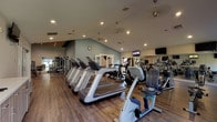 This single-floor fitness center is thoughtfully designed to accommodate a wide range of workout styles and fitness goals. The space is divided into several distinct zones, each purposefully outfitted to maximize functionality and comfort.

The main cardio area features rows of treadmills, stationary bikes, and elliptical machines arranged beneath vaulted ceilings with recessed lighting and pendant fixtures. Multiple wall-mounted flat-screen TVs are positioned throughout, and large windows along one wall invite natural light and outdoor views, creating an energizing atmosphere. Durable wood-style flooring runs throughout the facility, lending a clean, modern aesthetic.

An adjacent strength training zone includes cable machines, a multi-station functional trainer, adjustable benches, and a full rack of dumbbells. Floor-to-ceiling mirrors amplify the sense of space and support proper form. A distinctive slatted wood ceiling treatment adds a refined, contemporary design element to this area.

A dedicated functional training studio houses a comprehensive all-in-one rig stocked with medicine balls, kettlebells, battle ropes, resistance bands, and a heavy bag, offering versatile options for high-intensity and cross-training workouts.

A separate group exercise room provides an open, flexible space complete with stability balls, a step platform, yoga mats, a wall-mounted TV, and a smart fitness display, making it ideal for guided classes or solo stretching sessions.
