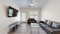 This single-story space offers a clean, modern aesthetic throughout, with light-toned wood-style flooring and crisp white walls providing a consistent, cohesive look. The living area is generously sized, accommodating multiple seating arrangements including a full sofa and loveseat. Natural light filters in through windows fitted with plantation shutters, while a ceiling fan overhead aids in climate comfort. A wall-mounted flat-screen TV and media console complete the functional setup. A glass-top coffee table anchors the space, and a private exterior door provides convenient outdoor access.

Each bedroom follows a consistent layout, featuring built-in wall shelving above a dedicated desk area with a chair, a dresser, a bed frame, and a window with blinds. Ceiling fans are present in select bedrooms, and each room benefits from access to a closet.

The kitchen is outfitted with white shaker-style cabinetry, light quartz countertops, and stainless steel appliances including a range, over-the-range microwave, and sink. An adjacent louvered-door closet houses in-unit washer and dryer connections. The bathroom features a full tub-shower combination with brass-toned fixtures set against white walls and wood-style flooring.