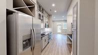 This single-floor unit offers a well-planned layout that maximizes every square foot of its compact footprint. The combined kitchen and living area serves as the heart of the space, showcasing warm hardwood-style flooring, neutral greige walls, and abundant natural light flowing in through multiple windows fitted with blinds. A glass-paneled entry door with transom window further brightens the living area, creating an inviting, airy atmosphere.

The kitchen is thoughtfully appointed with gray shaker-style cabinetry accented by matte black hardware, dark countertops, and a textured white tile backsplash. Stainless steel appliances — including a French door refrigerator with ice dispenser, electric range, built-in microwave, and dishwasher — provide a fully equipped culinary space. A built-in bench nook adjacent to the kitchen adds a practical mudroom-style touch.

Both bathrooms are finished to a high standard. One features floor-to-ceiling marble-look tile in a spacious glass-enclosed walk-in shower with a built-in bench. The other offers a white vanity with dark countertops, marble-style tile flooring, and matte black hardware throughout.

A dedicated walk-in closet provides generous built-in shelving and hanging rods, offering ample storage. Recessed lighting throughout the space ensures consistent, modern illumination in every room.