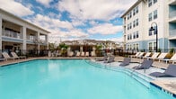 Located at 4027 Watercraft Ferry Ave in Wilmington, North Carolina, this outdoor amenity space is designed to offer a resort-inspired experience within a vibrant residential community. The centerpiece is a generously sized swimming pool with a sparkling turquoise finish, surrounded by a spacious pool deck furnished with sleek lounge chairs, ideal for relaxing under the coastal Carolina sky. A dedicated pool house structure anchors the poolside area, providing a functional and attractive focal point for the space.

A covered pavilion with white columned supports and light blue lap siding serves as a shaded gathering area, equipped with an outdoor television and seating—perfect for socializing or catching a game outdoors. The pavilion opens directly onto the surrounding courtyard, where a prominent round fire pit anchors a paved social area, creating an inviting atmosphere for evening gatherings.

Meticulously maintained landscaping features mulched garden beds, ornamental grasses, and young trees that add greenery and visual interest throughout the grounds. A black wrought-iron perimeter fence provides a clean boundary around the pool area while maintaining an open, airy feel. The surrounding multi-story residential buildings, finished in soft blue and white hues, frame the space and contribute to the community's cohesive coastal architectural aesthetic.