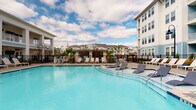 Located at 4027 Watercraft Ferry Ave in Wilmington, North Carolina, this outdoor amenity space is designed to offer a resort-inspired experience within a vibrant residential community. The centerpiece is a generously sized swimming pool with a sparkling turquoise finish, surrounded by a spacious pool deck furnished with sleek lounge chairs, ideal for relaxing under the coastal Carolina sky. A dedicated pool house structure anchors the poolside area, providing a functional and attractive focal point for the space.

A covered pavilion with white columned supports and light blue lap siding serves as a shaded gathering area, equipped with an outdoor television and seating—perfect for socializing or catching a game outdoors. The pavilion opens directly onto the surrounding courtyard, where a prominent round fire pit anchors a paved social area, creating an inviting atmosphere for evening gatherings.

Meticulously maintained landscaping features mulched garden beds, ornamental grasses, and young trees that add greenery and visual interest throughout the grounds. A black wrought-iron perimeter fence provides a clean boundary around the pool area while maintaining an open, airy feel. The surrounding multi-story residential buildings, finished in soft blue and white hues, frame the space and contribute to the community's cohesive coastal architectural aesthetic.