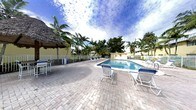 This outdoor pool area embodies a relaxed, tropical ambiance characteristic of Florida living. The centerpiece is a well-maintained rectangular swimming pool with clear blue water, framed by a generously sized paver brick deck that provides ample space for lounging and socializing. Multiple lounge chairs and seating arrangements are positioned around the pool, offering plenty of spots to relax and soak up the sun.

A standout feature of this space is the thatched-roof tiki bar structure positioned along the poolside, adding a resort-like, tropical character to the setting. The area is enclosed by a low fence, ensuring a sense of privacy and safety while maintaining an open, airy feel. Towering palm trees and lush tropical greenery surround the perimeter, creating a natural canopy that enhances the lush, laid-back atmosphere.

The surrounding buildings, finished in a warm yellow exterior, complement the tropical aesthetic. The broad open sky above, paired with the mature landscaping, gives the space a bright, expansive feel ideal for outdoor relaxation. Overall, the pool area presents a welcoming, resort-inspired retreat designed for leisure.