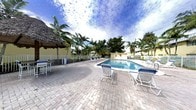 This outdoor pool area embodies a relaxed, tropical ambiance characteristic of Florida living. The centerpiece is a well-maintained rectangular swimming pool with clear blue water, framed by a generously sized paver brick deck that provides ample space for lounging and socializing. Multiple lounge chairs and seating arrangements are positioned around the pool, offering plenty of spots to relax and soak up the sun.

A standout feature of this space is the thatched-roof tiki bar structure positioned along the poolside, adding a resort-like, tropical character to the setting. The area is enclosed by a low fence, ensuring a sense of privacy and safety while maintaining an open, airy feel. Towering palm trees and lush tropical greenery surround the perimeter, creating a natural canopy that enhances the lush, laid-back atmosphere.

The surrounding buildings, finished in a warm yellow exterior, complement the tropical aesthetic. The broad open sky above, paired with the mature landscaping, gives the space a bright, expansive feel ideal for outdoor relaxation. Overall, the pool area presents a welcoming, resort-inspired retreat designed for leisure.
