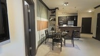 This single-floor space offers a thoughtfully designed open-concept layout that seamlessly connects the dining area and kitchen, creating a cohesive and functional living environment. The dining area is anchored by a round glass-top table paired with elegant black chairs, positioned beneath tasteful pendant and track lighting that casts a warm, inviting glow throughout the space. A striking large-scale wall mural featuring abstract botanical motifs in sage green, warm orange, and neutral tones serves as a bold focal point, adding depth and artistic character to the room. Decorative open shelving nearby displays carefully curated accessories, enhancing the overall aesthetic. The adjacent kitchen is outfitted with dark cabinetry, stainless steel appliances, and a breakfast bar counter, combining practicality with a modern sensibility. Overhead track lighting illuminates the kitchen workspace efficiently. The flooring features light-toned tile with subtle circular detailing, contributing to a clean and polished foundation throughout. The overall ambiance of the space is contemporary and refined, with a cohesive dark-and-light contrast palette, layered lighting, and decorative accents that work together to create a welcoming and visually engaging interior.