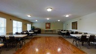 This single-floor community or event hall is designed to accommodate a variety of social gatherings and activities. The space is anchored by a generously sized, polished hardwood dance floor that occupies the central portion of the room, providing a warm and inviting focal point. Surrounding the dance floor, multiple folding tables and chairs are arranged along the perimeter, offering ample seating for guests during events or dining occasions.

The room is illuminated by a combination of flush-mount ceiling light fixtures and a track lighting system, ensuring even and functional lighting throughout. The textured ceiling adds a classic touch to the overall interior. Light-colored walls contribute to a bright and open ambiance, while a decorative vintage-style poster serves as a notable visual accent on the back wall.

Along one side of the room, a piano or organ is visible, suggesting the space is well-suited for live music performances. A large mirror panel is mounted on one wall, visually expanding the room. Natural light filters in through a glass door toward the front of the space, complementing the interior lighting. The layout is flexible and practical, making it suitable for dances, community events, meetings, or social gatherings.