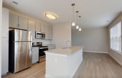 Located at 9755 Mill Centre Dr in Owings Mills, Maryland, this single-floor residence offers a thoughtfully designed open-concept layout with contemporary finishes throughout. The kitchen serves as the heart of the home, featuring a large granite-topped island with an integrated sink, gray shaker-style cabinetry, stainless steel appliances including a refrigerator, range, and over-the-range microwave, and light wood-style flooring that extends seamlessly into the adjoining living and dining areas. Elegant pendant lighting hangs above the island, complementing the flush-mount ceiling fixtures throughout. The living and dining area benefits from abundant natural light, with multiple windows dressed in horizontal blinds offering views of the surrounding greenery. The bedroom is finished with plush carpeting and a generously sized window that continues the theme of natural light. A hallway features mirrored sliding closet doors, maximizing both storage and the sense of space. The bathrooms are clean and functional, with one featuring a full tub-shower combination with white tile surround, a floating vanity, and light wood-style flooring. The neutral warm-beige color palette used throughout creates a cohesive and calming ambiance well-suited for modern living.