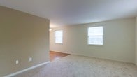 This single-floor residence offers a clean, straightforward layout designed for comfortable everyday living. The main living area is finished with soft, neutral-toned carpeting that extends throughout, creating a warm and cohesive feel underfoot. Multiple double-hung windows are positioned to maximize natural light, brightening the interior with an airy, open ambiance. The walls are painted in a soft, warm neutral tone that complements the light palette of the space.

An adjacent area — visible through an open passage — transitions to hardwood flooring, providing a functional and visually distinct zone that could serve as a dining area or foyer. A ceiling-mounted light fixture in this connected space adds a practical overhead lighting element. Clean white baseboards run throughout, adding a crisp finishing detail to the rooms.

The overall design is minimal and uncluttered, making the space feel open and versatile. The layout flows naturally between the carpeted and hard-floored zones, offering flexibility for a variety of furniture arrangements and uses. The consistent neutral color scheme throughout the space ensures a unified, calm atmosphere that is well-suited for a range of interior styles and personal preferences.