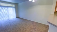 This single-floor space presents a clean, open living area finished with wall-to-wall carpeting in a neutral tone, contributing to a quiet and comfortable atmosphere. The room is characterized by smooth, light-colored walls trimmed with white baseboards, creating a bright and simple aesthetic throughout. A ceiling-mounted light fixture provides overhead illumination, supplementing the natural light that enters through a sliding glass door fitted with vertical blinds along the left wall. The sliding door serves as both a source of daylight and a potential access point to an outdoor area. Electrical outlets are positioned along the walls, offering practical functionality across the space. The overall layout is straightforward and versatile, with no obstructions limiting how the room can be arranged or utilized. The neutral palette and carpeted flooring lend the space a understated, move-in-ready character suitable for a variety of uses.