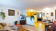This single-floor residence offers a functional and connected layout, with living, kitchen, and bedroom areas flowing naturally into one another. The living room serves as the main gathering space, featuring carpeted flooring, a textured ceiling, and neutral walls that create a warm, understated backdrop. A television and wooden media console anchor one side of the room, while a cushioned ottoman and area rug add comfort and definition to the seating area. Houseplants placed throughout contribute a lively, homey touch.

A half-wall partition separates the living room from the kitchen, creating a sense of openness while maintaining distinct zones. The kitchen features warm honey-toned cabinetry that extends to the upper walls, providing ample storage. A full-sized refrigerator and overhead lighting complete the functional kitchen setup. The transition from carpeted living space to hardwood-style flooring marks the shift into the kitchen and adjoining areas.

Visible through a doorway, the bedroom offers a glimpse of natural light filtering through a window with curtains, hardwood-style flooring, and enough space for essential furnishings. Overall, the residence presents a cozy and practical living environment with a classic, homey character.